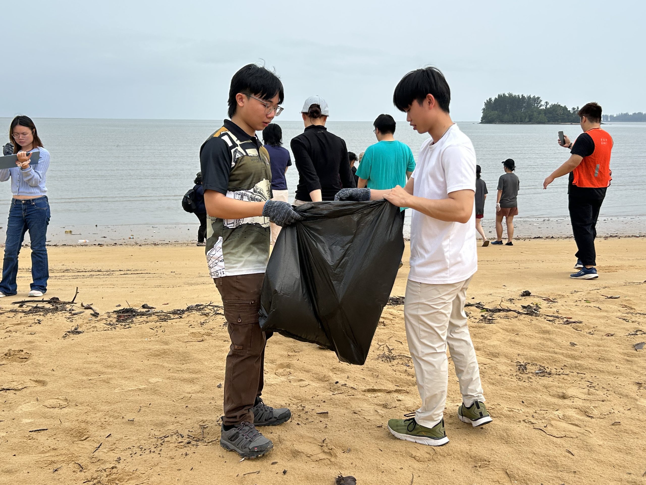 Two young volunteers in white shirts stand on the sand at Pasir Panjang beach, one holding open a large black garbage bag while others walk along the shoreline behind them.